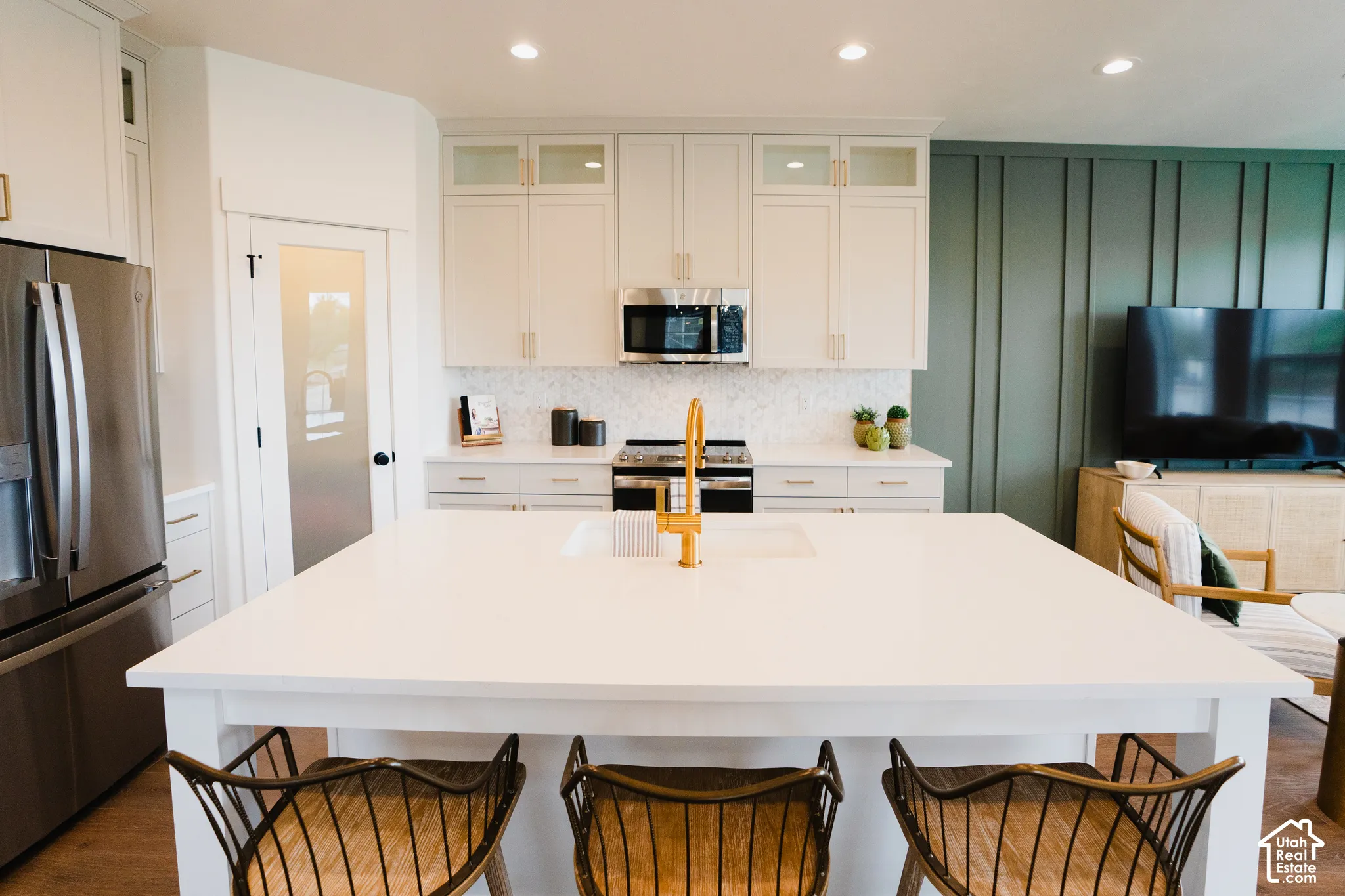 Expansive kitchen island with a built-in sink and gold-tone faucet, white shaker-style cabinetry with gold-tone hardware, stainless steel appliances, and wood-finish flooring