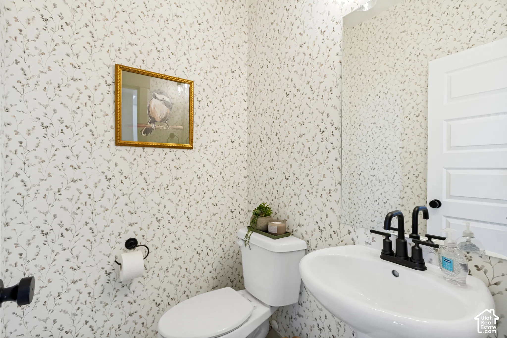 Pedestal sink with a matte black faucet, wall-mounted mirror, and patterned wallpaper