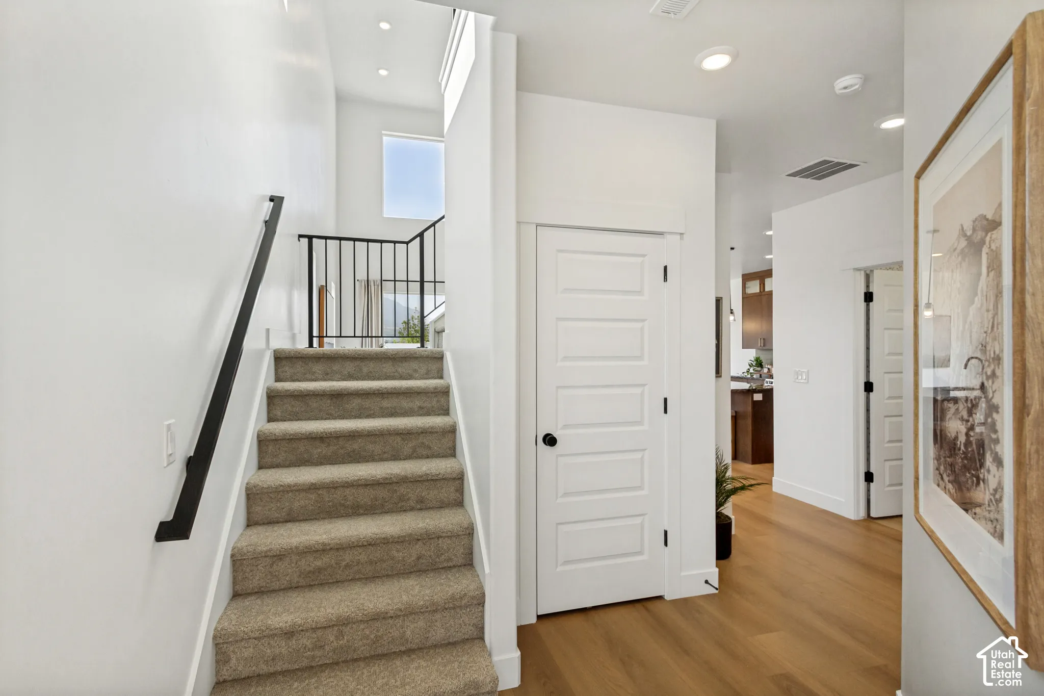 Carpeted staircase with black metal railing, five-panel interior door, recessed lighting, and wood-finish flooring