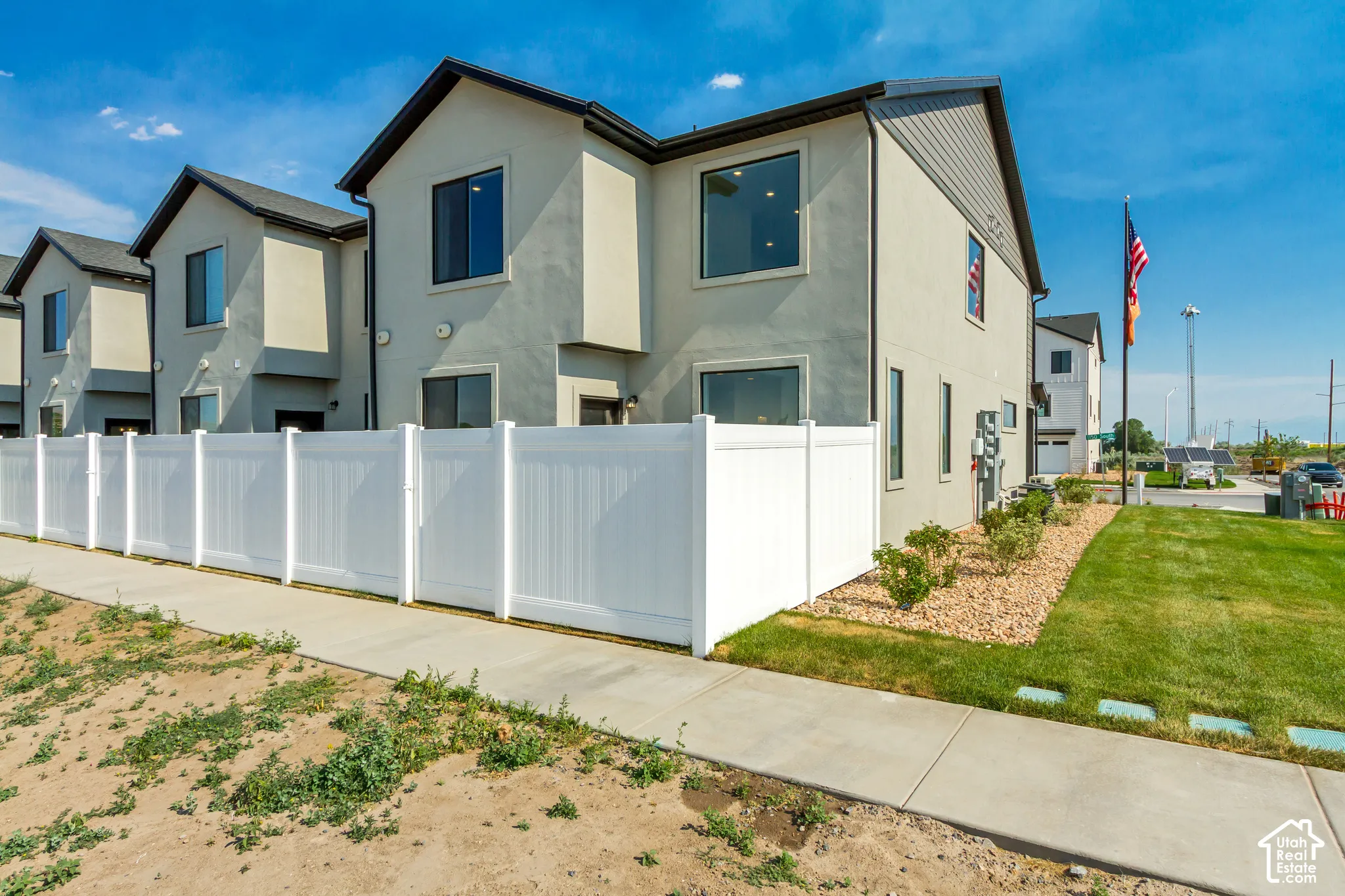 Stucco exterior with gabled roofline, multiple windows, white privacy fencing, manicured lawn, and landscaped rock beds
