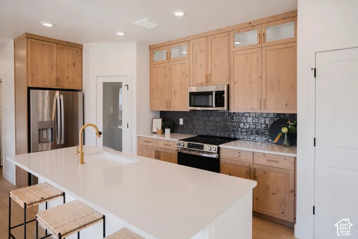 Kitchen featuring light wood-finish cabinetry, stainless steel appliances, a white solid surface island with an undermount sink, and a dark tile backsplash