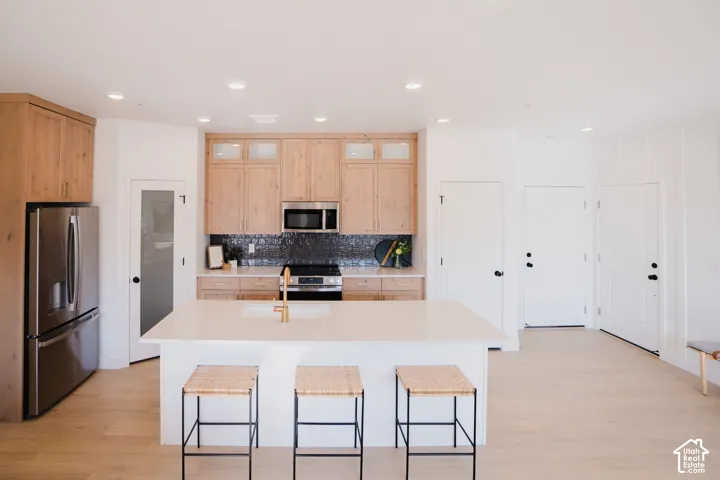 Kitchen featuring a large central island, stainless steel appliances, light wood-finish cabinetry, and a dark tile backsplash