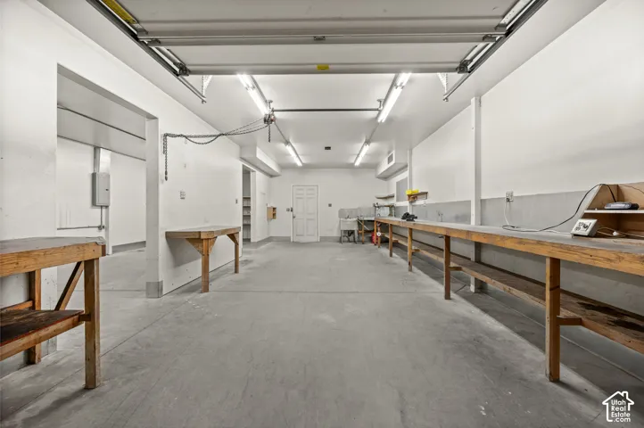 Concrete flooring and exposed overhead garage door track in a spacious utility area featuring multiple built-in wooden workbenches and overhead fluorescent lighting