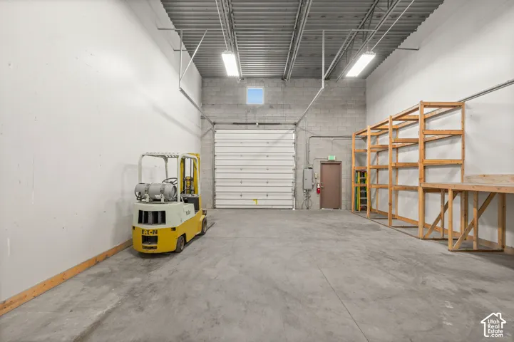 Spacious industrial interior featuring a roll-up door, concrete block wall construction, exposed metal ceiling, and built-in wood shelving with a workbench