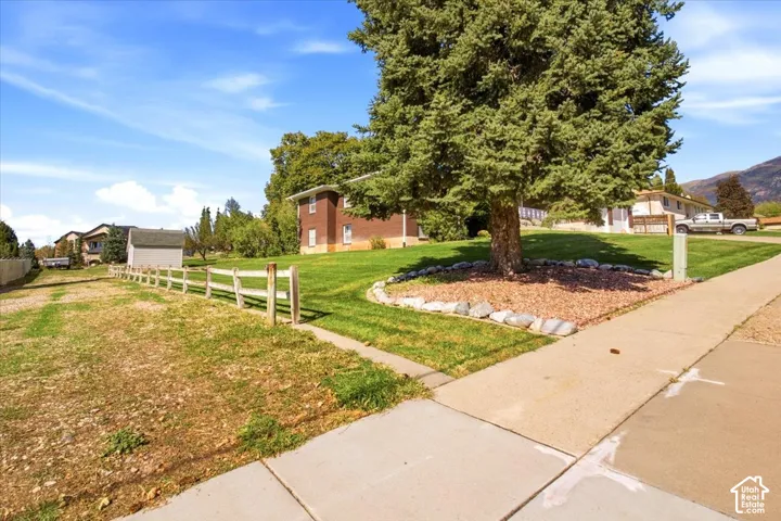 Expansive lot featuring a substantial mature tree, a split-rail fence, a paved sidewalk, and a manicured lawn