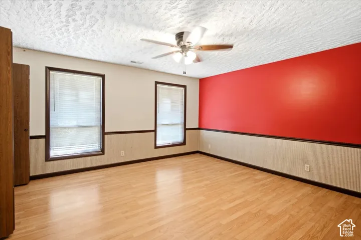 Spacious room featuring wood-finish flooring, two large windows with black frames and white blinds, ceiling fan with light fixture, and a striking red accent wall paired with neutral beige wainscoting and upper walls