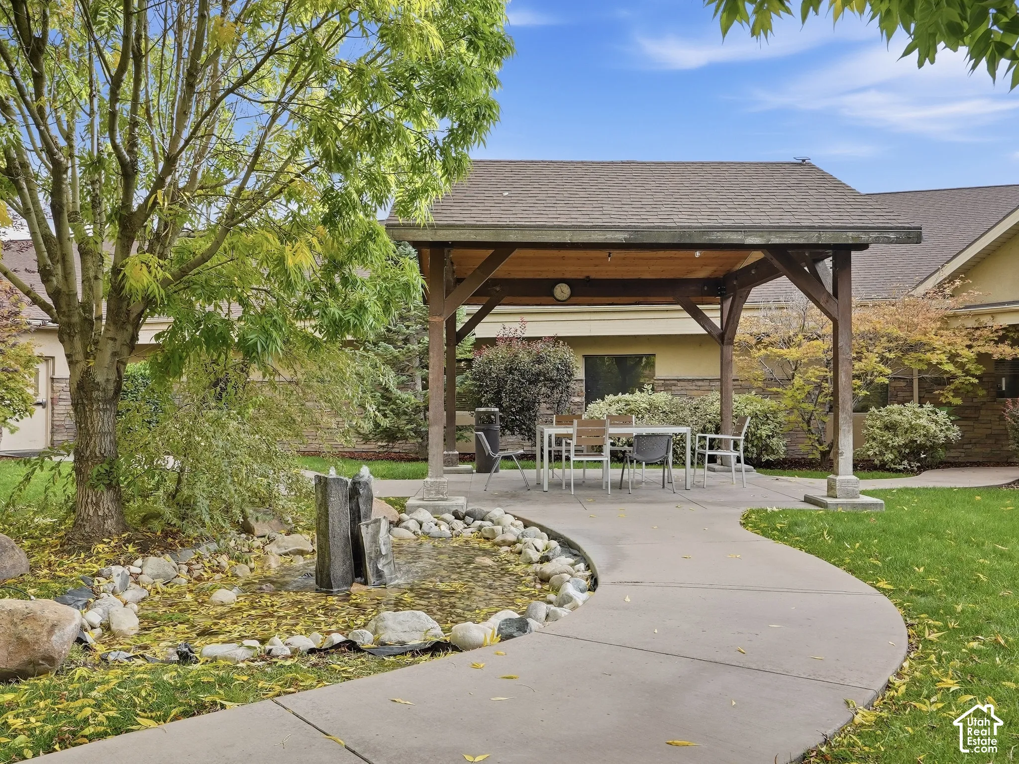 Covered patio structure with a concrete pad, a rock-lined pond with a water feature, and a paved walkway