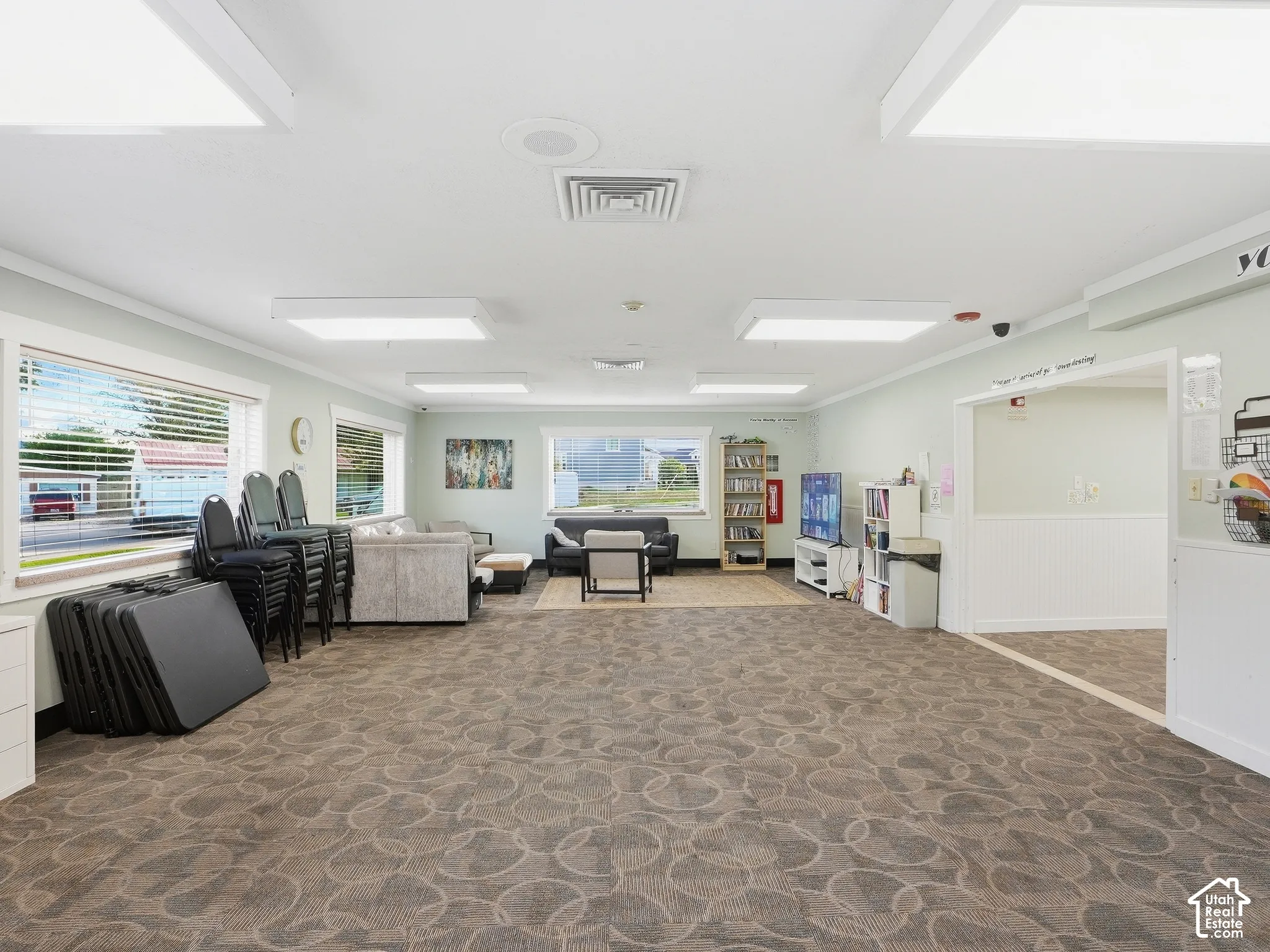 Spacious common area featuring patterned carpeting, multiple windows with blinds, recessed lighting, and an entryway with wainscoting