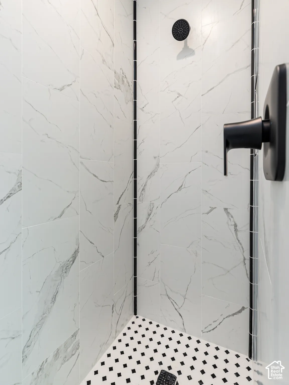 White tile shower with black accent trim, matte black showerhead, matte black shower control, and black and white mosaic tile flooring