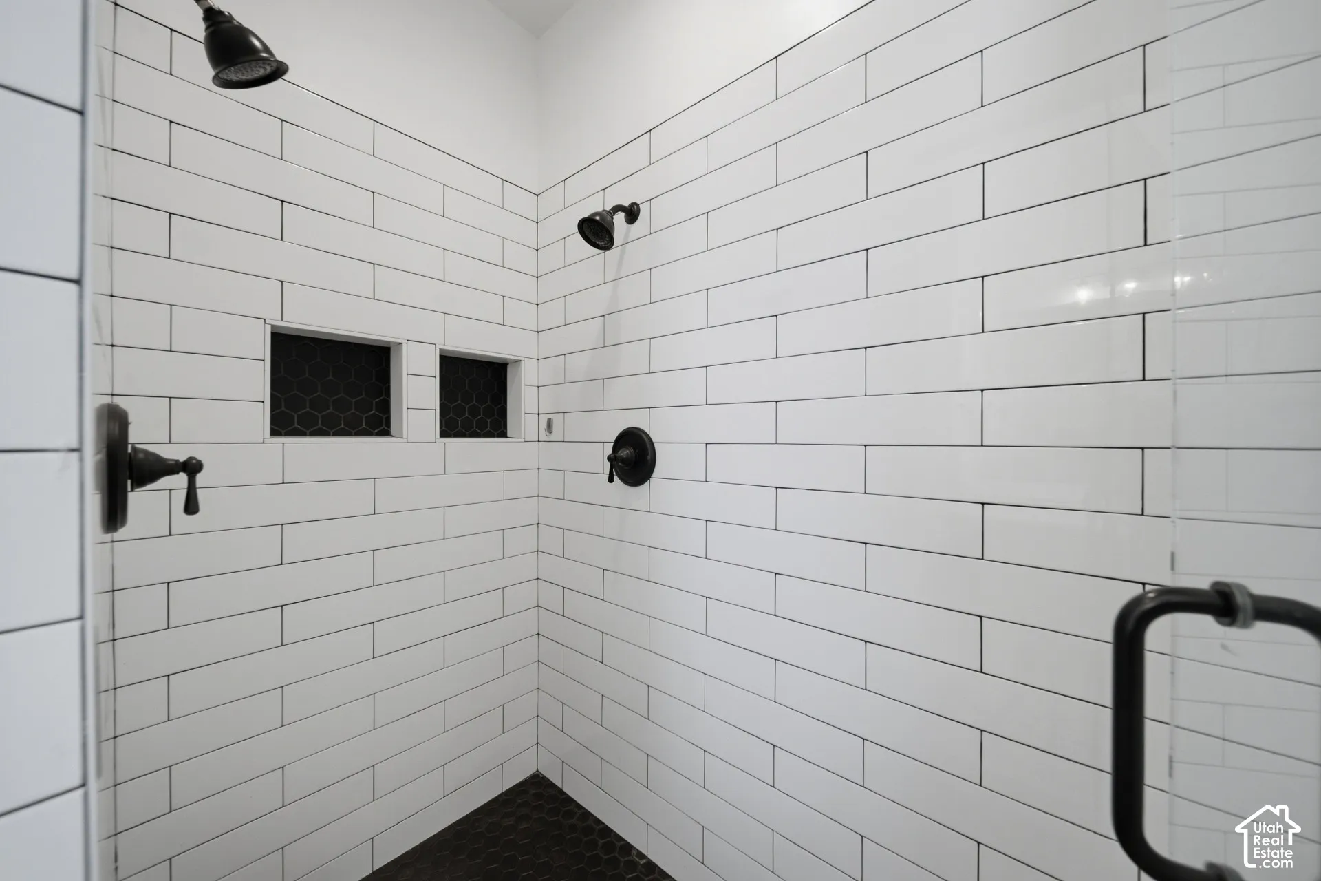 Shower with white subway tile, black hexagon tile flooring, dual shower heads, recessed shelving, and a clear glass enclosure