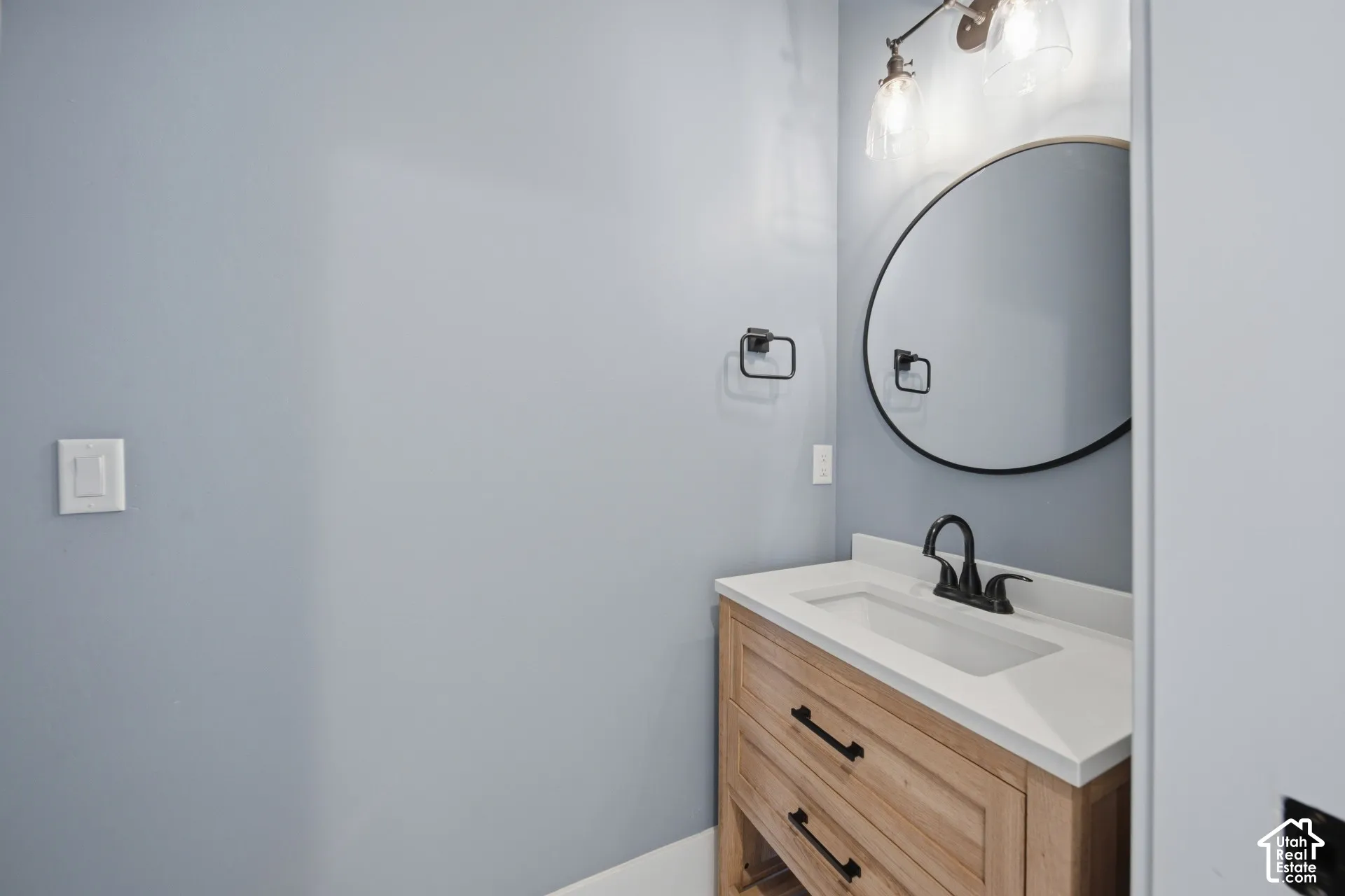 Wood-finish vanity with drawer storage, white countertop with integrated sink, matte black faucet, round wall-mounted mirror, and wall-mounted light fixture