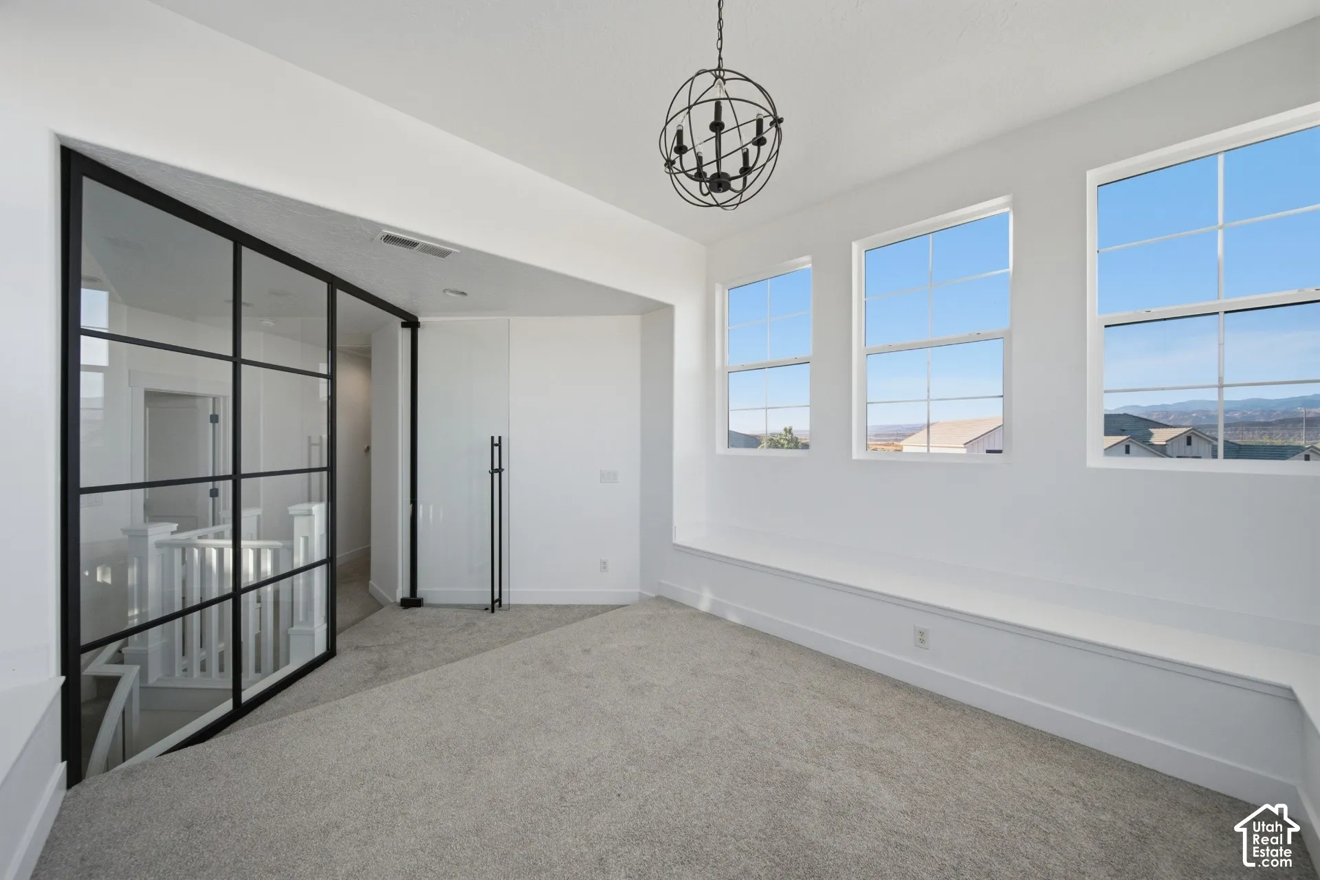 Carpeted room featuring a spherical chandelier, three large windows, a window bench, and a pivoting glass panel wall with black framing