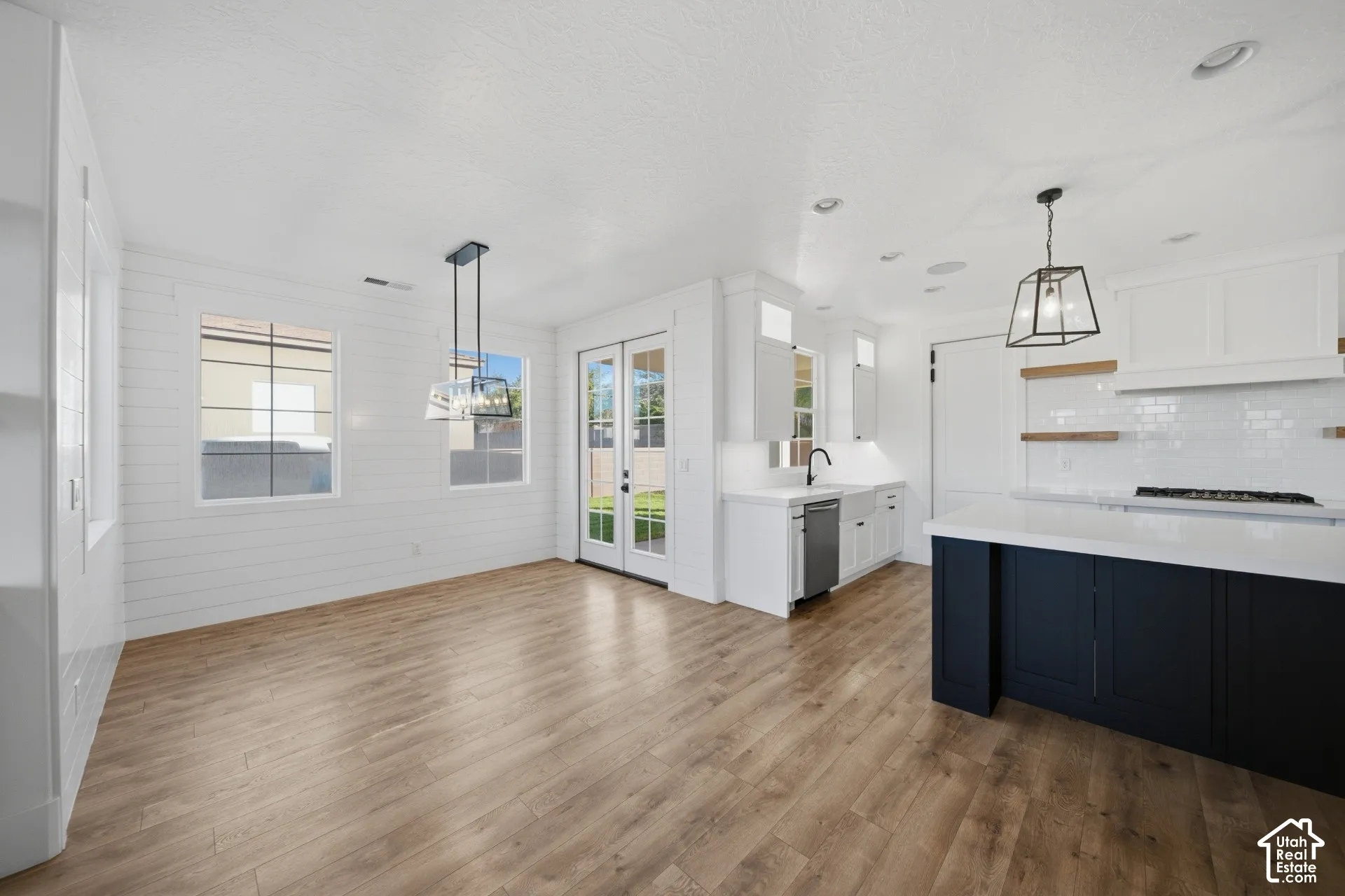 Open-concept living space with wood-finish flooring, white shiplap walls, pendant lighting, and a kitchen featuring white cabinetry, a dark island, and a tile backsplash