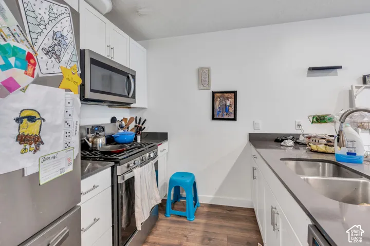 White cabinetry with brushed nickel hardware, stainless steel appliances, dark countertops, wood-finish flooring, and a double basin sink