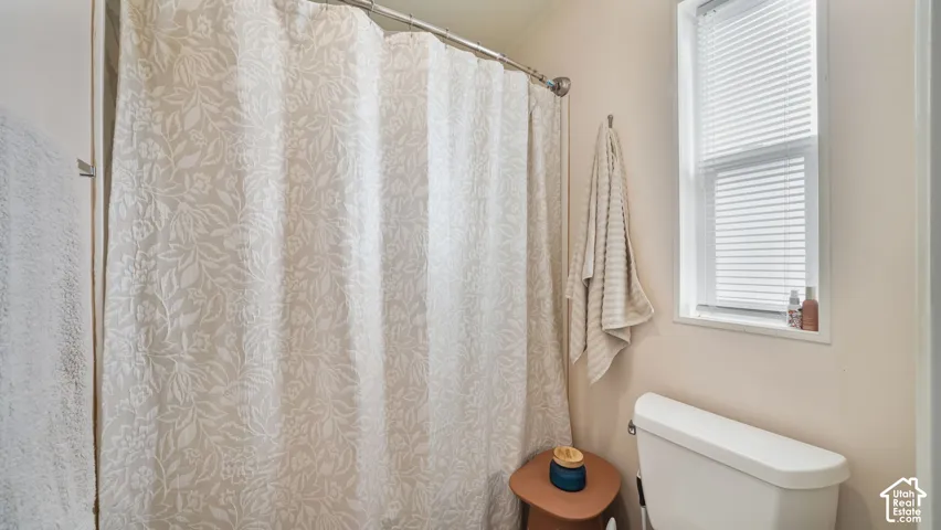 Bathroom featuring a patterned shower curtain with metal rod, white porcelain toilet, wooden accent table, and natural light from a window with plantation shutters
