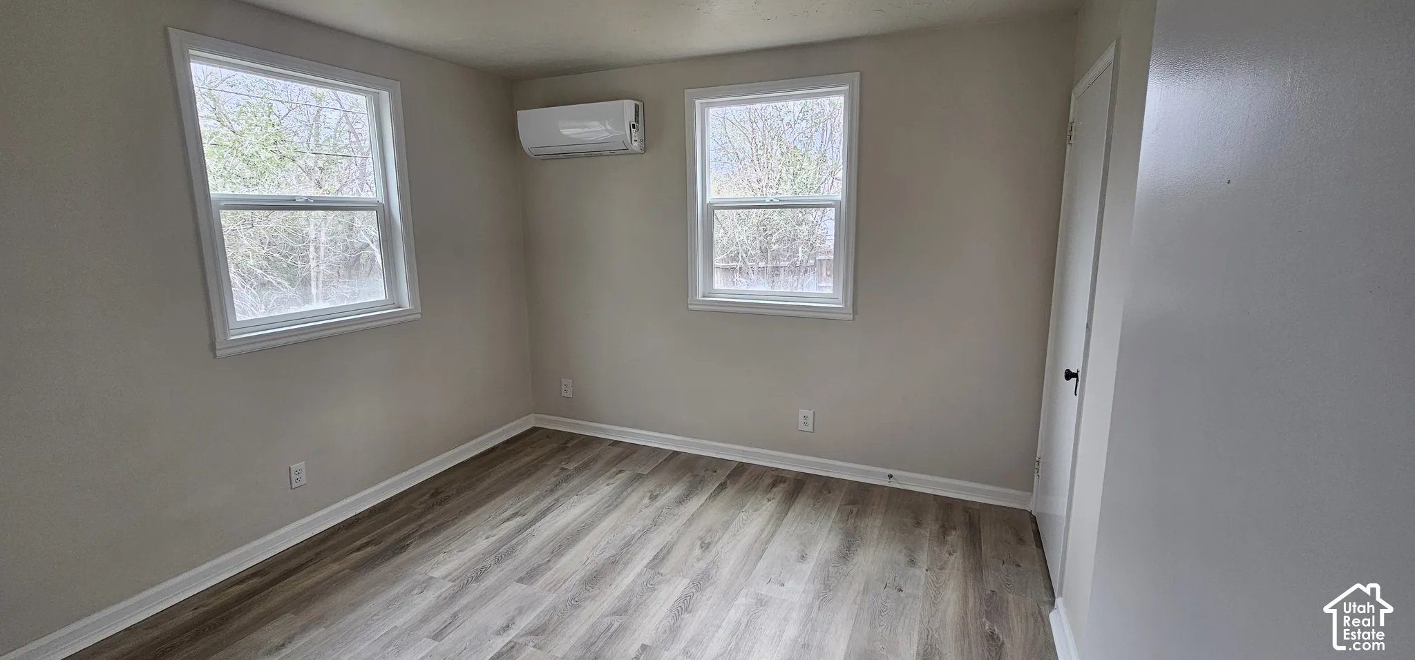 Light-toned wood-finish flooring, two large windows with white trim, and a wall-mounted air conditioning unit