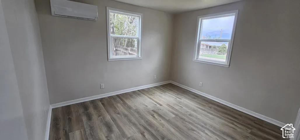 Wood-finish flooring, two windows with white trim, and a wall-mounted climate control unit