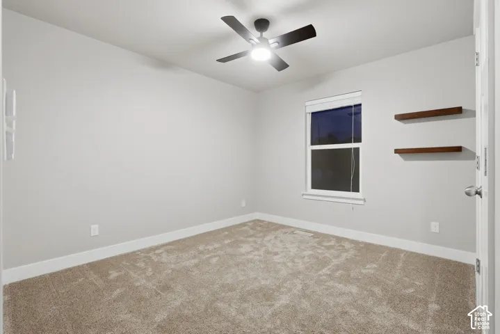 Neutral wall color, carpet flooring, window with blind, ceiling fan with integrated lighting, and two wall-mounted shelves