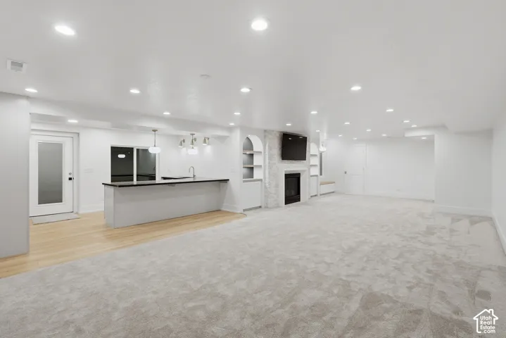 Recessed lighting illuminates the open-concept space featuring light wood-finish flooring, a kitchen island with dark countertops, and a white brick fireplace