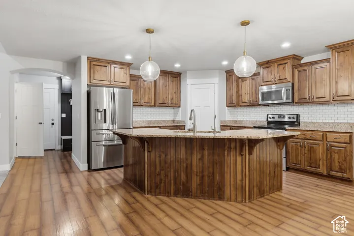 Kitchen featuring wood-finish flooring, wood cabinetry, stainless steel appliances, a center island with a sink, and pendant lighting