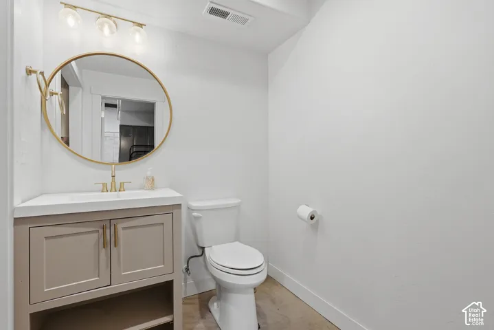 Powder room featuring a single vanity with a light gray cabinet, white countertop, and gold-finish fixtures including a circular mirror and three-light vanity bar