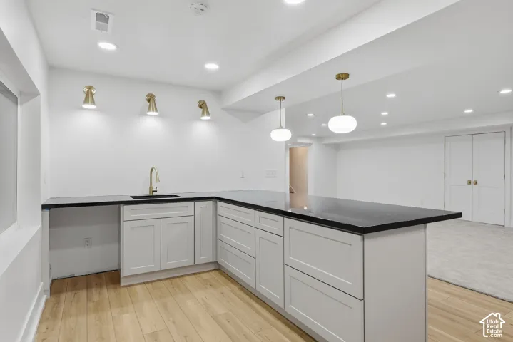 L-shaped countertop with an undermount sink and gold-finish faucet, light gray cabinetry, wood-finish flooring, recessed lighting, and wall-mounted sconces