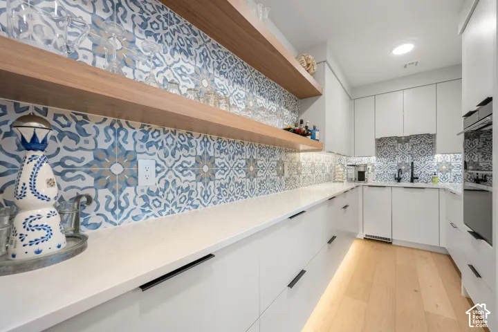 White flat-panel cabinetry with matte black hardware, white countertops, patterned tile backsplash, natural wood-finish open shelving, and light wood-finish flooring