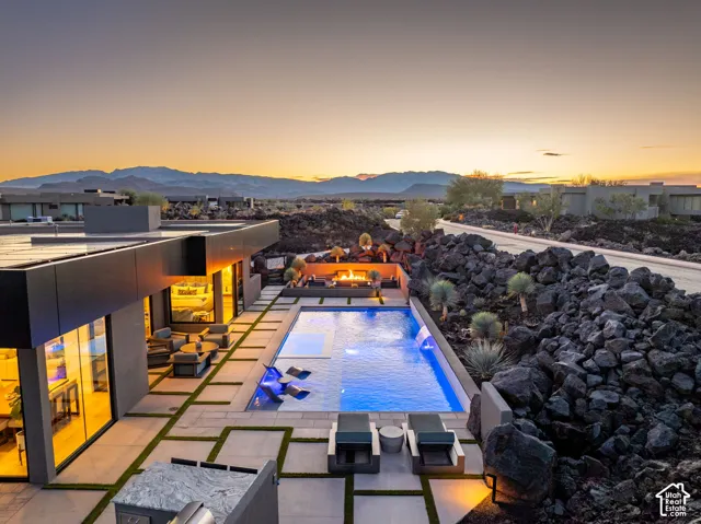 Modern architecture featuring a rectangular pool with integrated water features, a built-in outdoor kitchen with a grill, a fire pit, and expansive paving with artificial turf accents