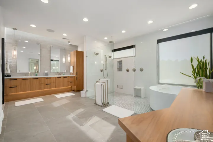 Dual vanity with wood-finish cabinetry, frameless glass shower enclosure, freestanding soaking tub, large format tile flooring, and recessed lighting