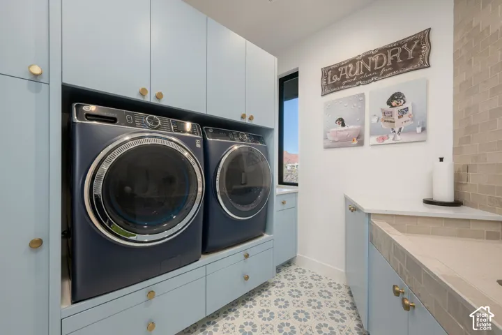 Built-in washer and dryer set, light blue cabinetry with gold-tone hardware, patterned tile flooring, and a utility sink with a tiled backsplash