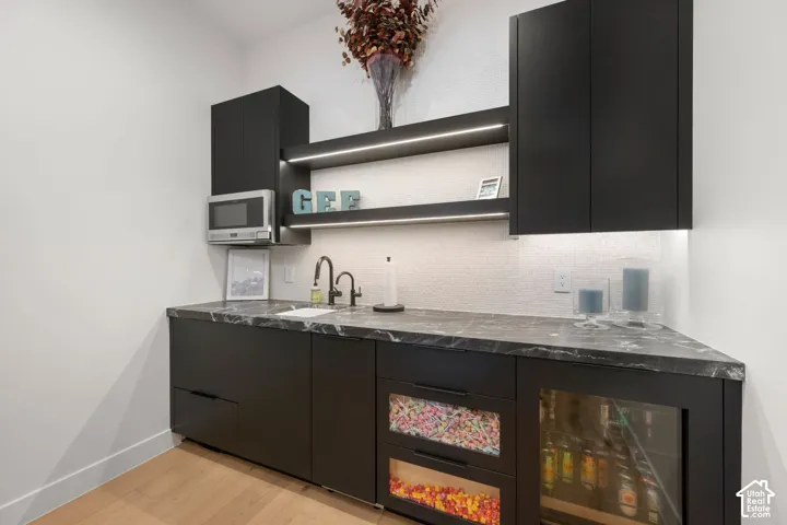 Built-in cabinetry with dark finishes, dark countertop, undermount sink with a gooseneck faucet, integrated beverage cooler, and open shelving with integrated lighting