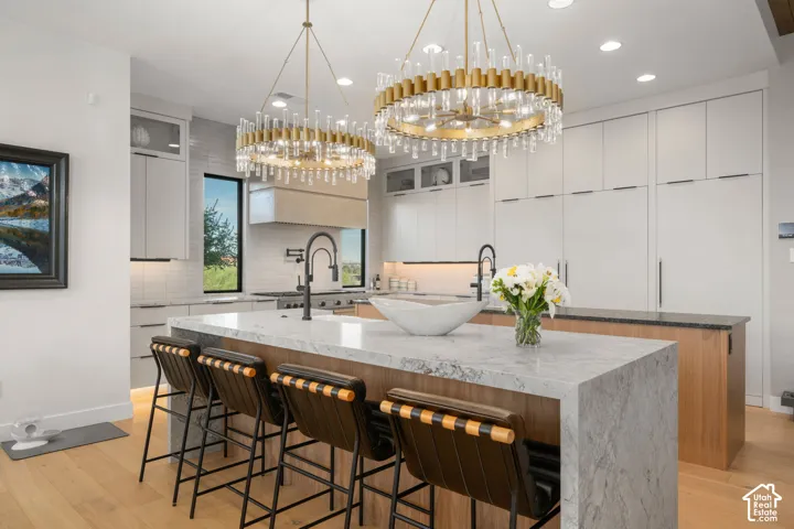 Kitchen featuring dual gold-finish chandeliers, a waterfall countertop island, wood-finish flooring, a white subway tile backsplash, and integrated paneled appliances
