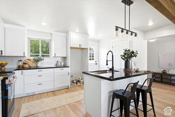 White cabinetry with dark hardware, black countertops, wood-finish flooring, island with sink and faucet, and a linear pendant light fixture