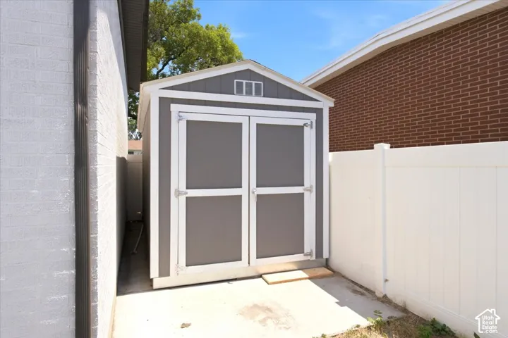 Gable roof storage shed with double doors, set on a concrete pad, bordered by a white privacy fence and brick-faced exterior