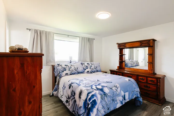 Bedroom featuring wood-finish flooring, wood-frame bed with blue patterned bedding, wood dresser with attached mirror, wood nightstand, recessed ceiling lighting, and large window with light curtains