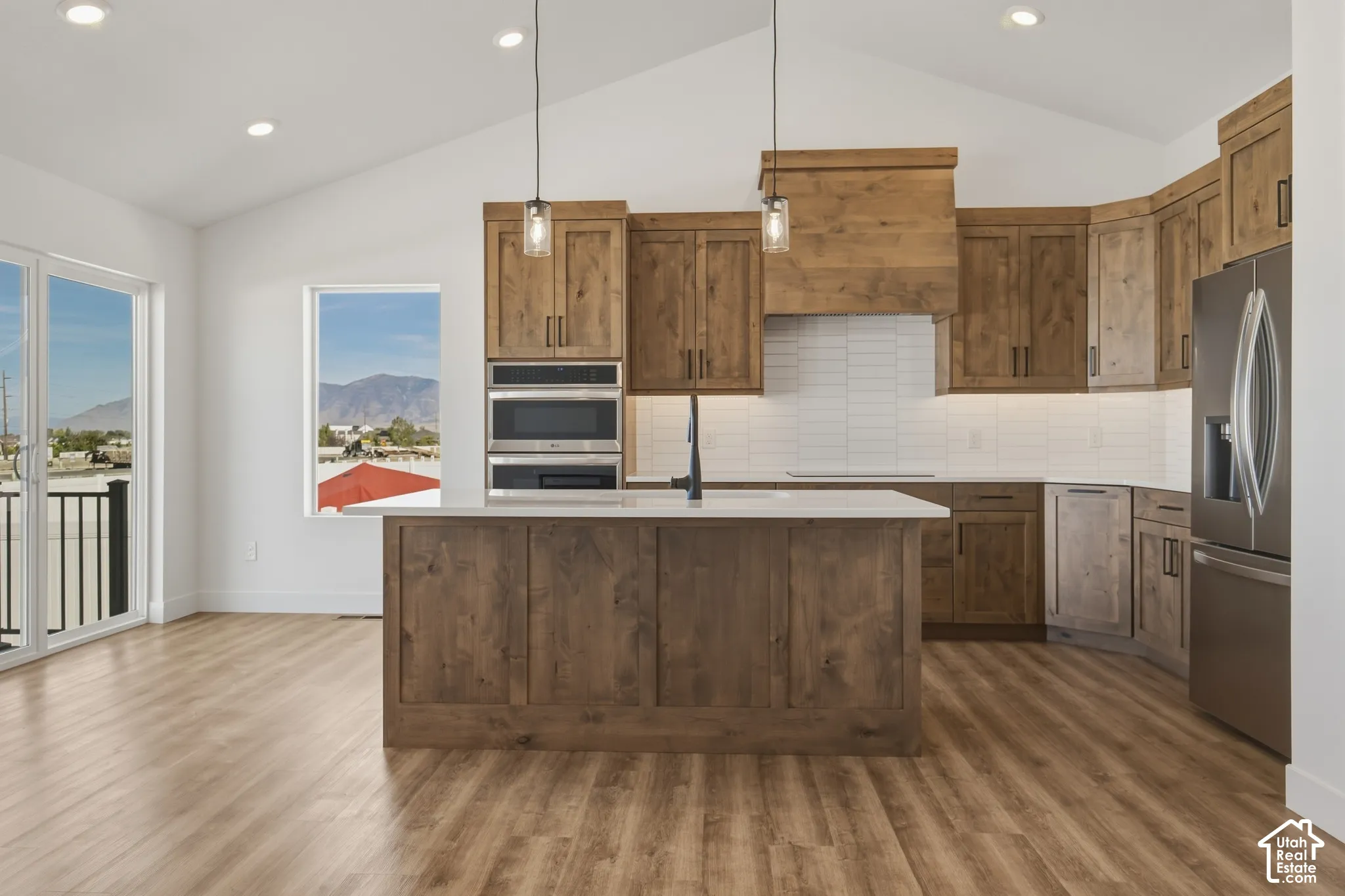 Vaulted ceiling kitchen featuring wood-finish cabinetry, a large kitchen island with a light-toned countertop, stainless steel appliances, and wood-finish flooring