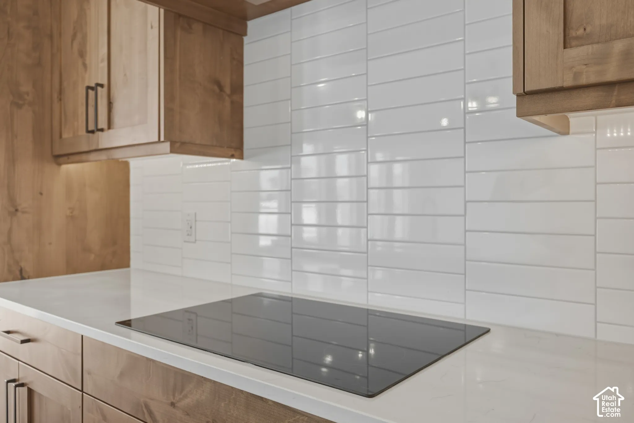 Wood-finish cabinetry with dark hardware, sleek cooktop, white countertop, white subway tile backsplash, and under-cabinet lighting