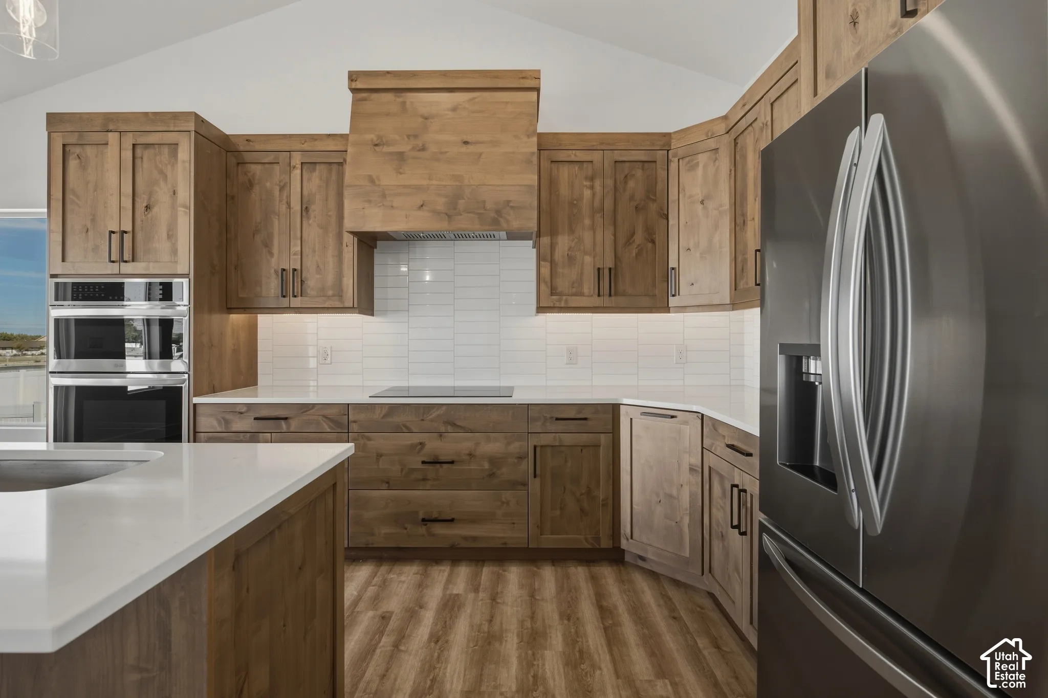 Vaulted ceiling with recessed lighting, wood-finish cabinetry, built-in double ovens, stainless steel refrigerator, and white subway tile backsplash