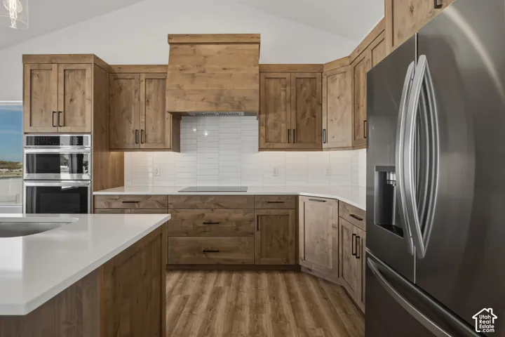 Vaulted ceiling with recessed lighting, wood-finish cabinetry, built-in double ovens, stainless steel refrigerator, and white subway tile backsplash