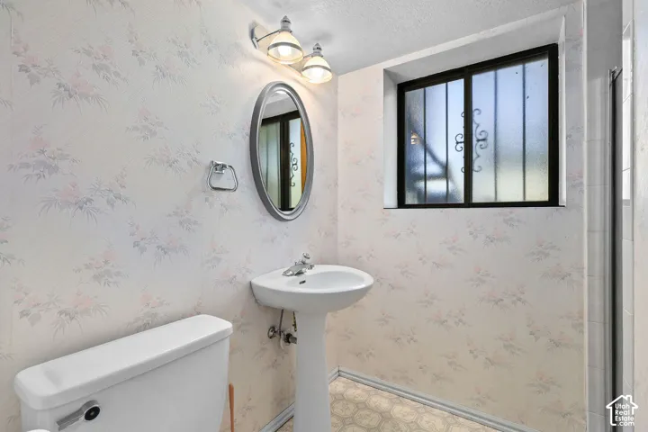 Pedestal sink with chrome faucet, oval mirror, wall-mounted light fixture, frosted window with security bars, and floral-patterned wallpaper
