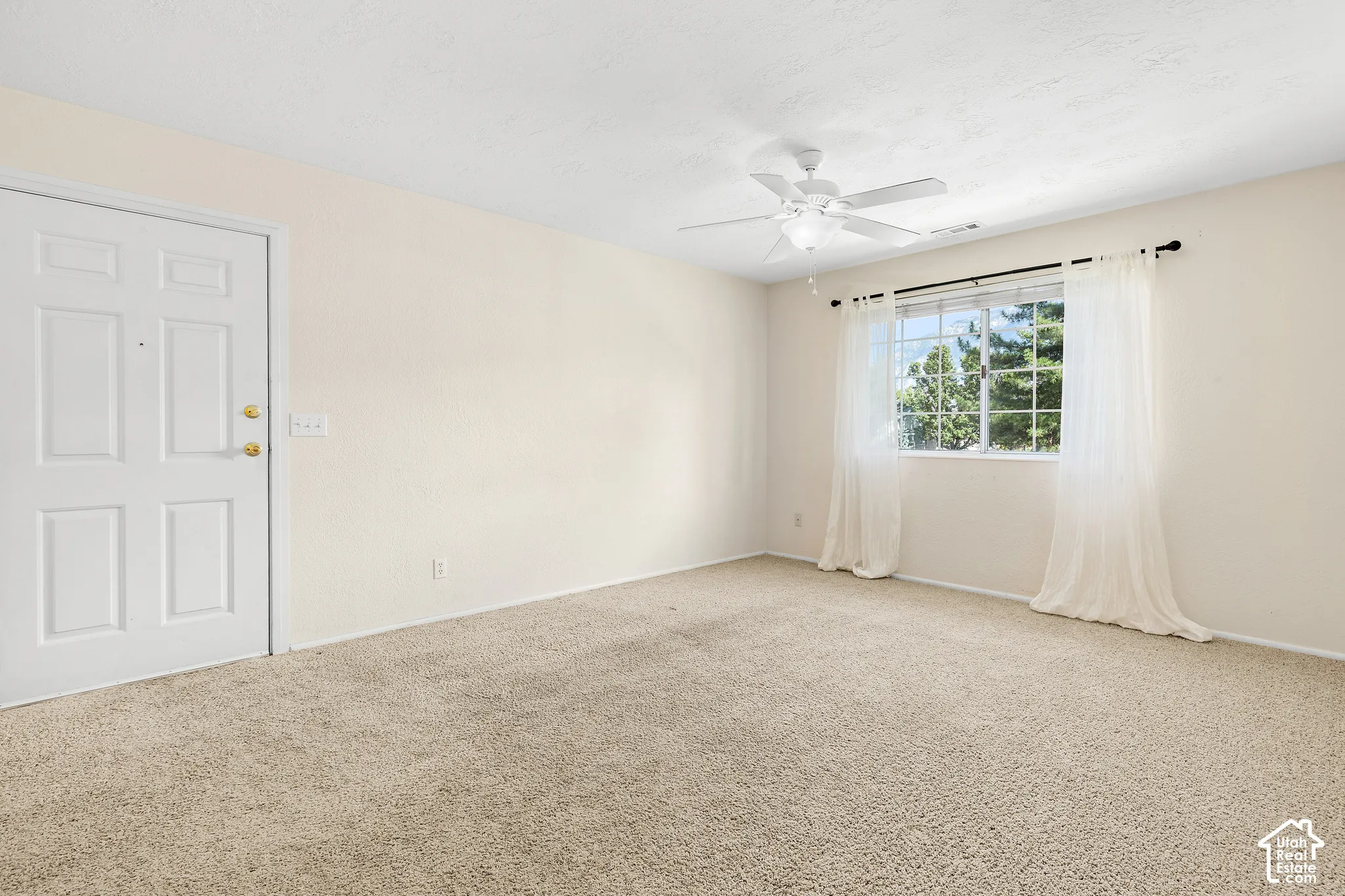 Textured carpet flooring, window with exterior scenery, paneled door with brass hardware, and a ceiling fan with integrated lighting