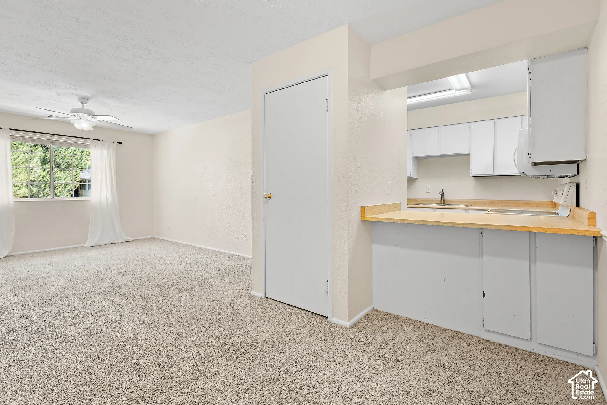 Carpeted living area with a ceiling fan, window, and pass-through to a kitchen featuring white cabinetry and light wood-finish countertops