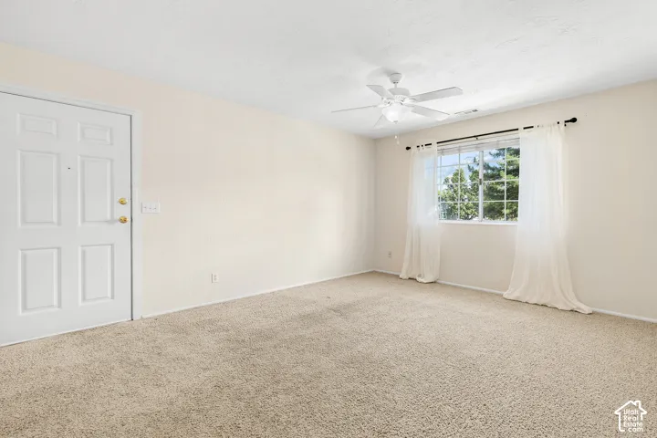 Textured carpet flooring, window with exterior scenery, paneled door with brass hardware, and a ceiling fan with integrated lighting