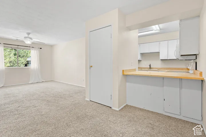 Carpeted living area with a ceiling fan, window, and pass-through to a kitchen featuring white cabinetry and light wood-finish countertops