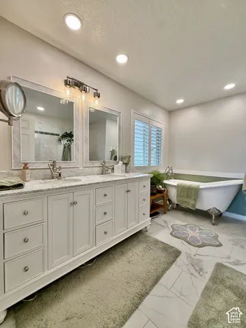 Dual sink vanity with stone countertop, freestanding clawfoot tub, recessed lighting, white louvered shutters, and large format floor tiling
