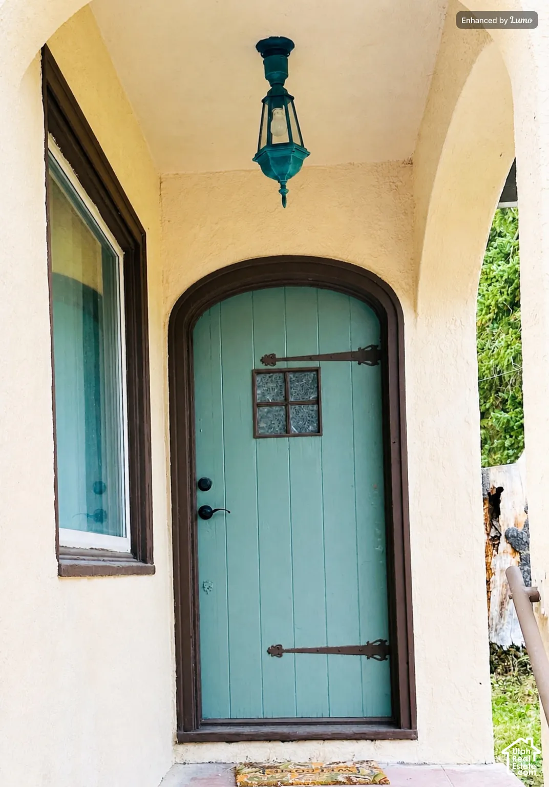 Arched entry featuring a teal door with decorative hardware, a small window with grilles, an overhead lantern, and stucco exterior