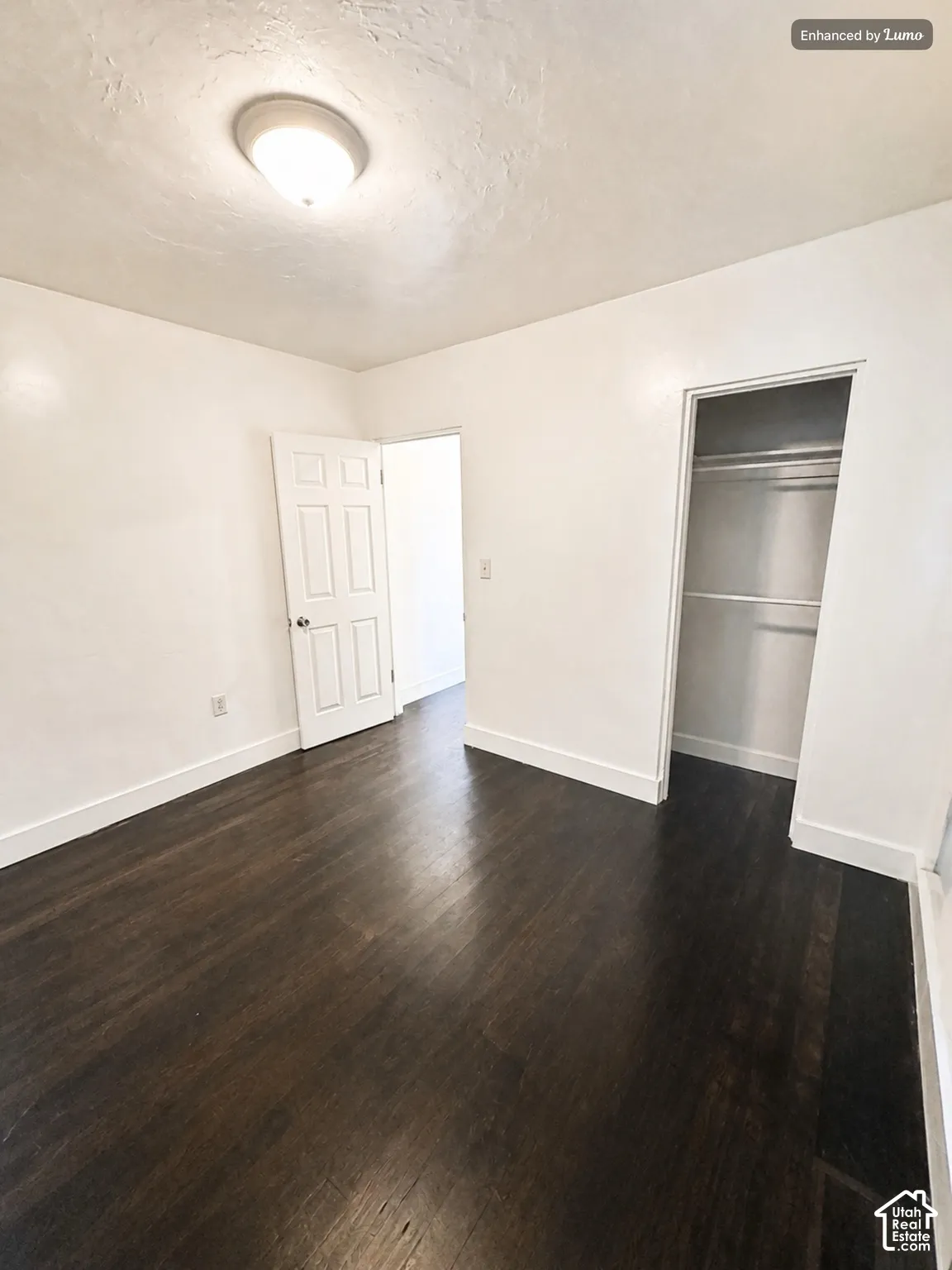 Dark wood-finish flooring, white painted walls, ceiling-mounted light fixture, white interior door, and built-in closet with shelving