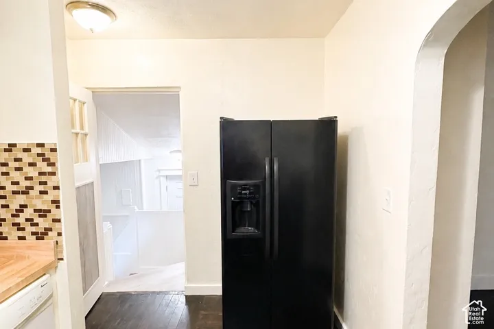 Black side-by-side refrigerator, multi-toned tile backsplash, wood-finish countertop, dark wood-finish flooring, and arched doorway