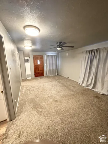 Spacious room featuring neutral carpeting, a dark wood-finish entry door, and a ceiling fan with integrated lighting