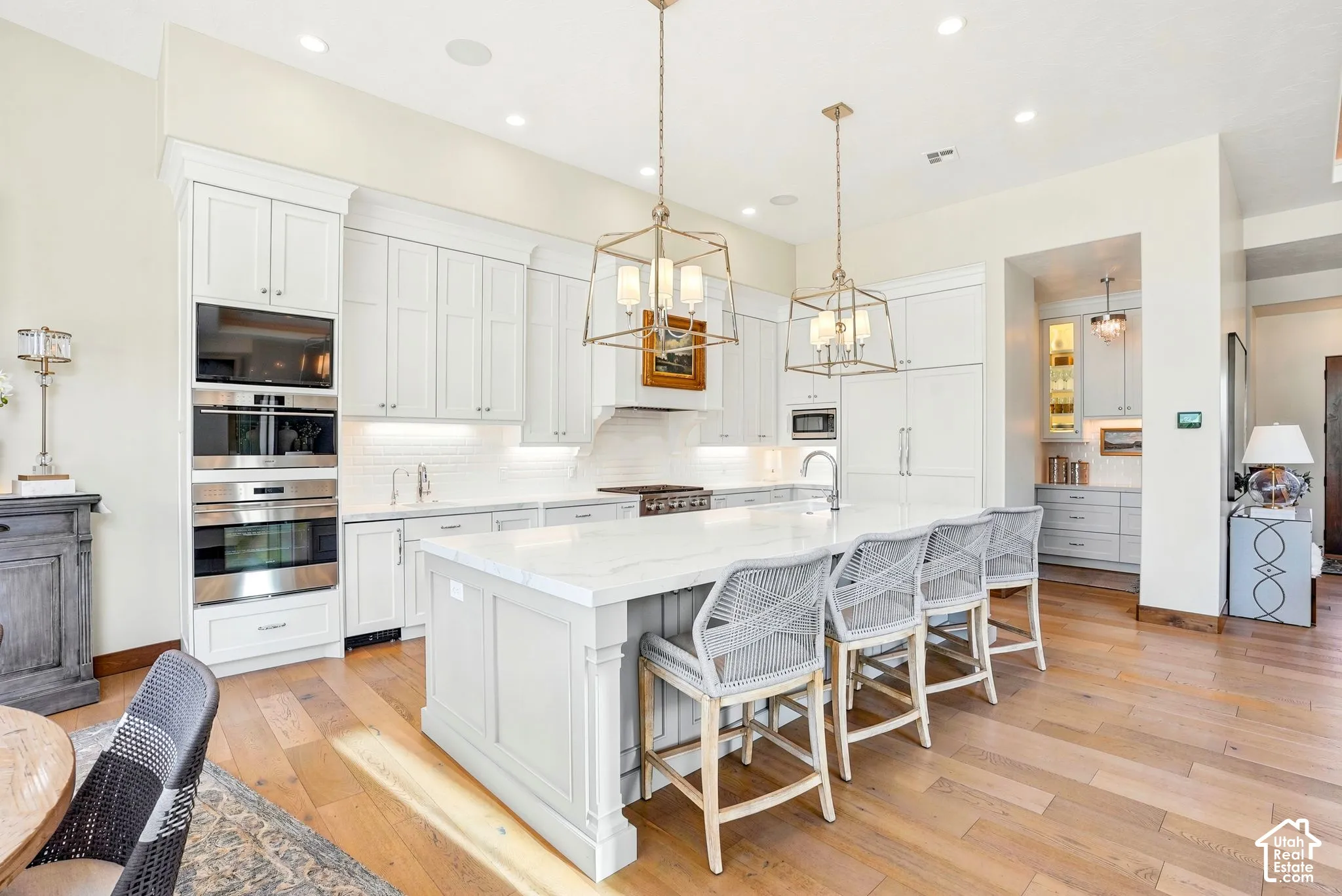 Custom white cabinetry, built-in stainless steel double ovens, large center island with countertop seating, wood-finish flooring, and recessed lighting